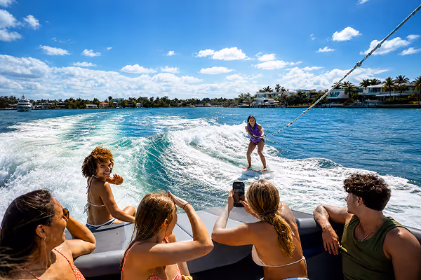 Young woman in a purple life vest wakesurfing on turquoise water with a city skyline and sunny sky in the background.