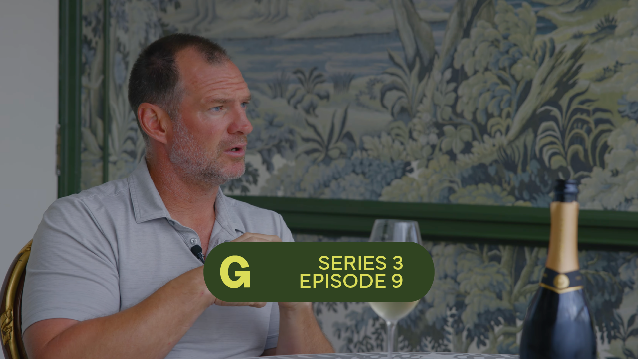 Man in gray shirt speaking while sitting at table with wine glass and bottle, floral wallpaper background.