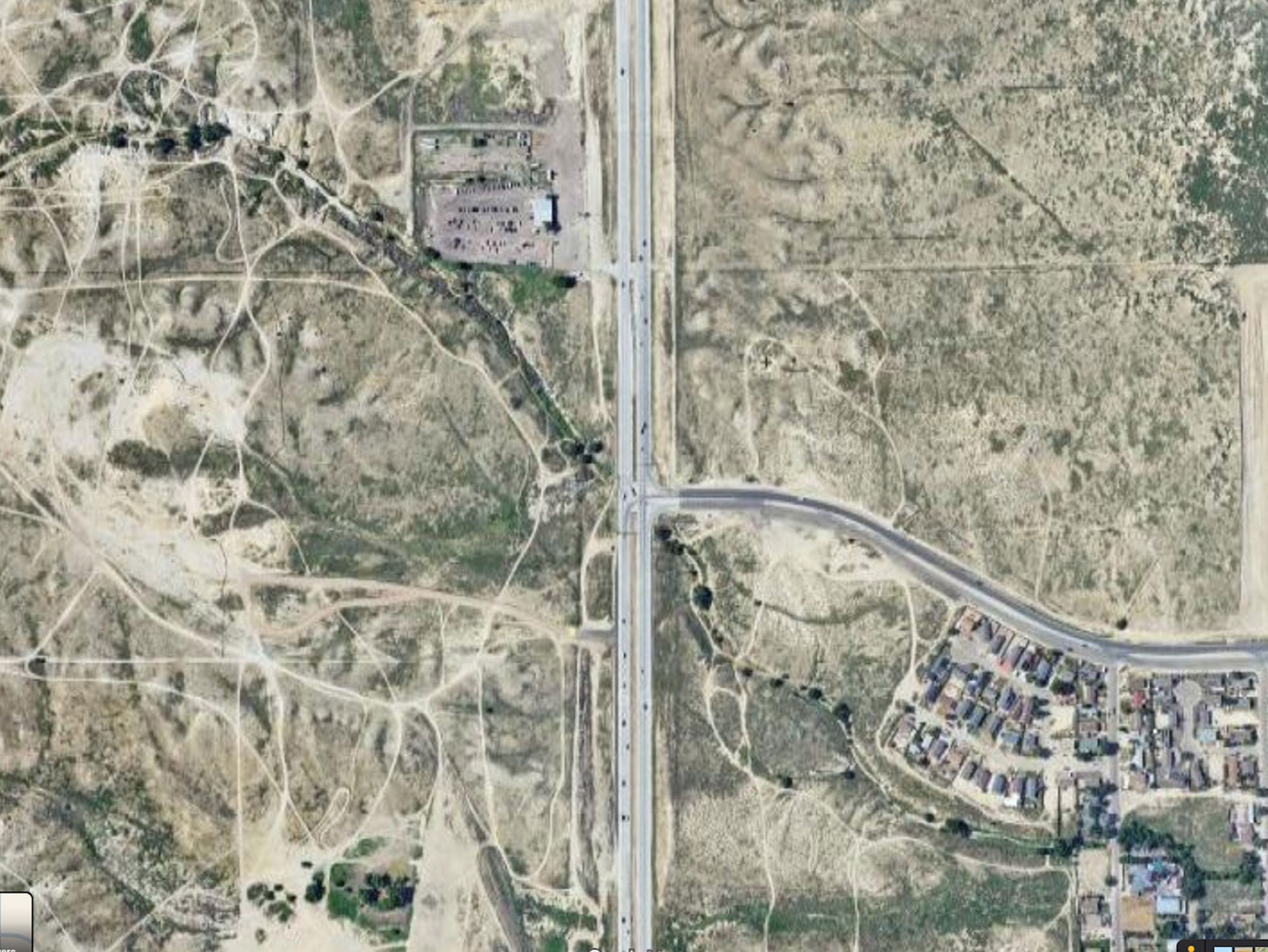 Aerial view of a highway intersecting a rural area with sparse vegetation, dirt paths, a small industrial facility, and a residential neighborhood.