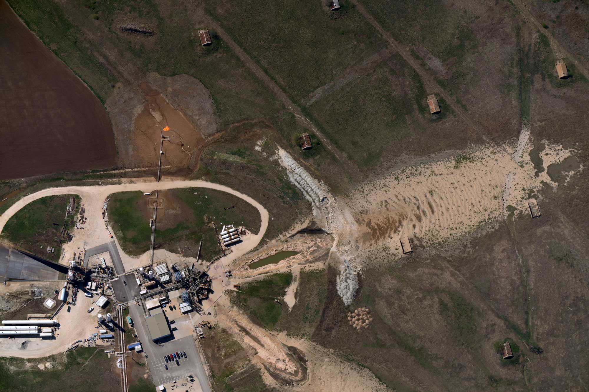 Aerial view of an industrial facility with equipment, pipes, and a parking area surrounded by open land with dirt and grass patches.