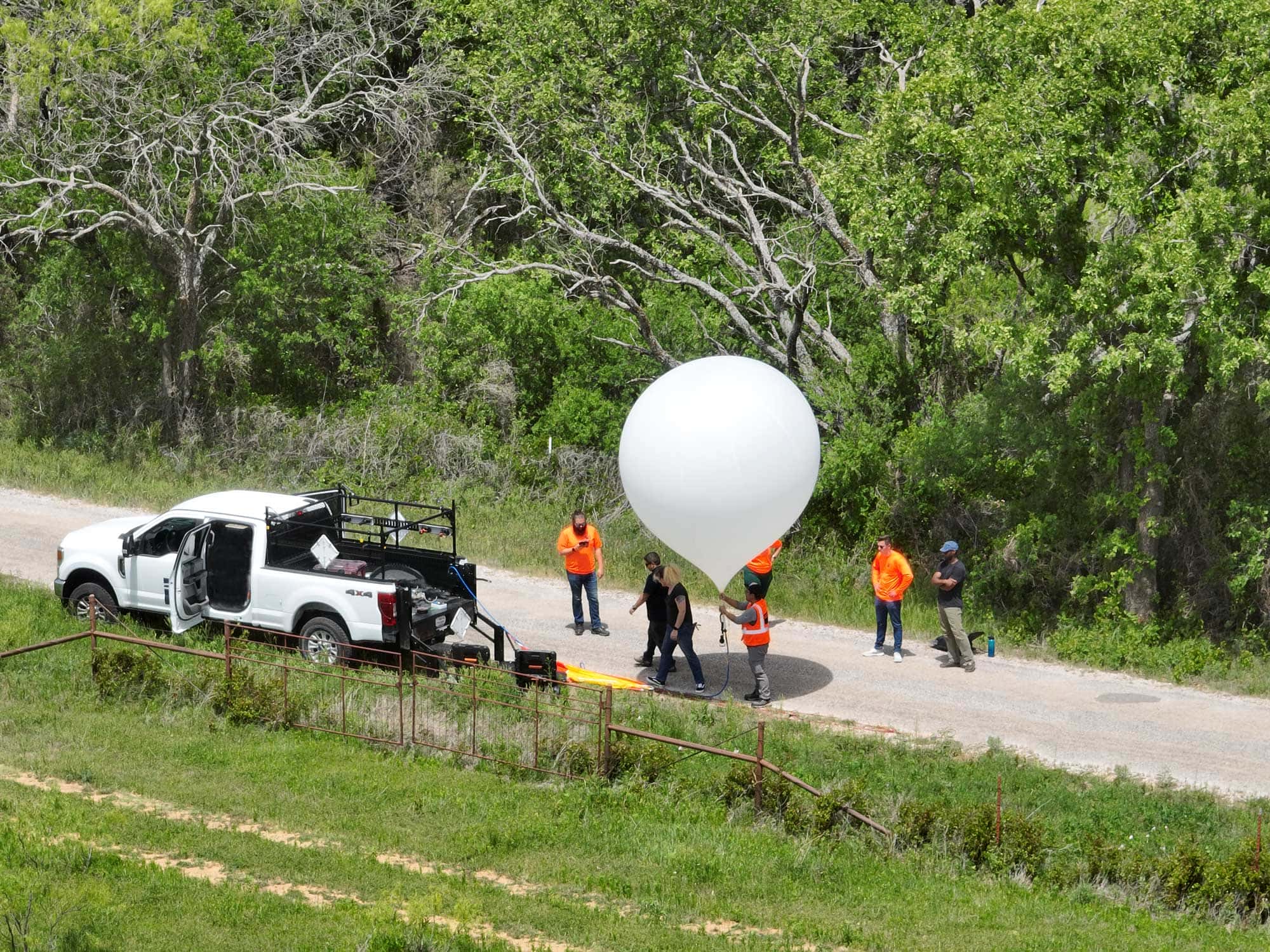 Group of people in safety vests and orange shirts preparing a large white weather balloon next to a white pickup truck on a gravel road surrounded by trees.