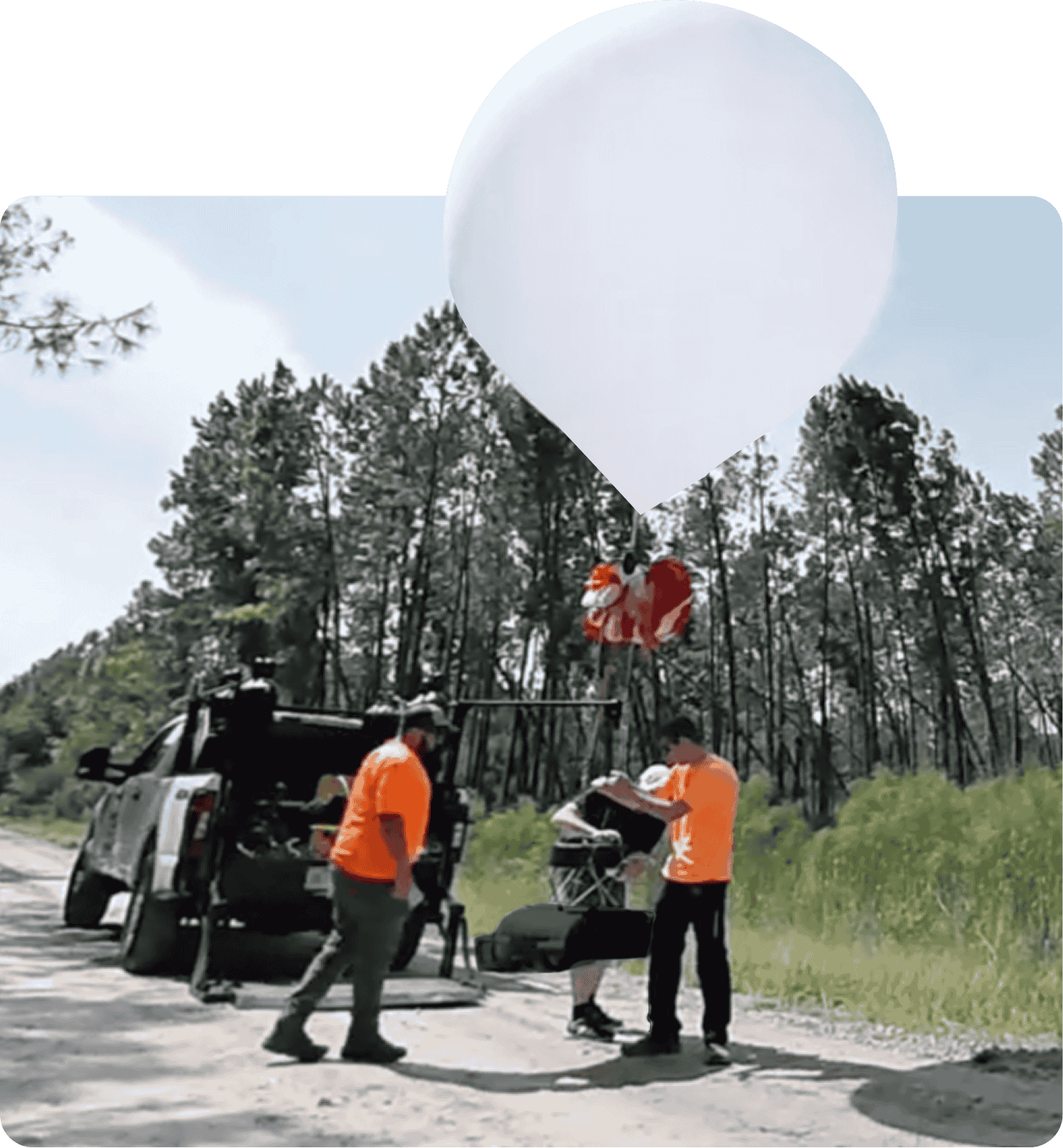 Three people in orange shirts preparing a large white weather balloon on a dirt road near a parked truck and forested area.