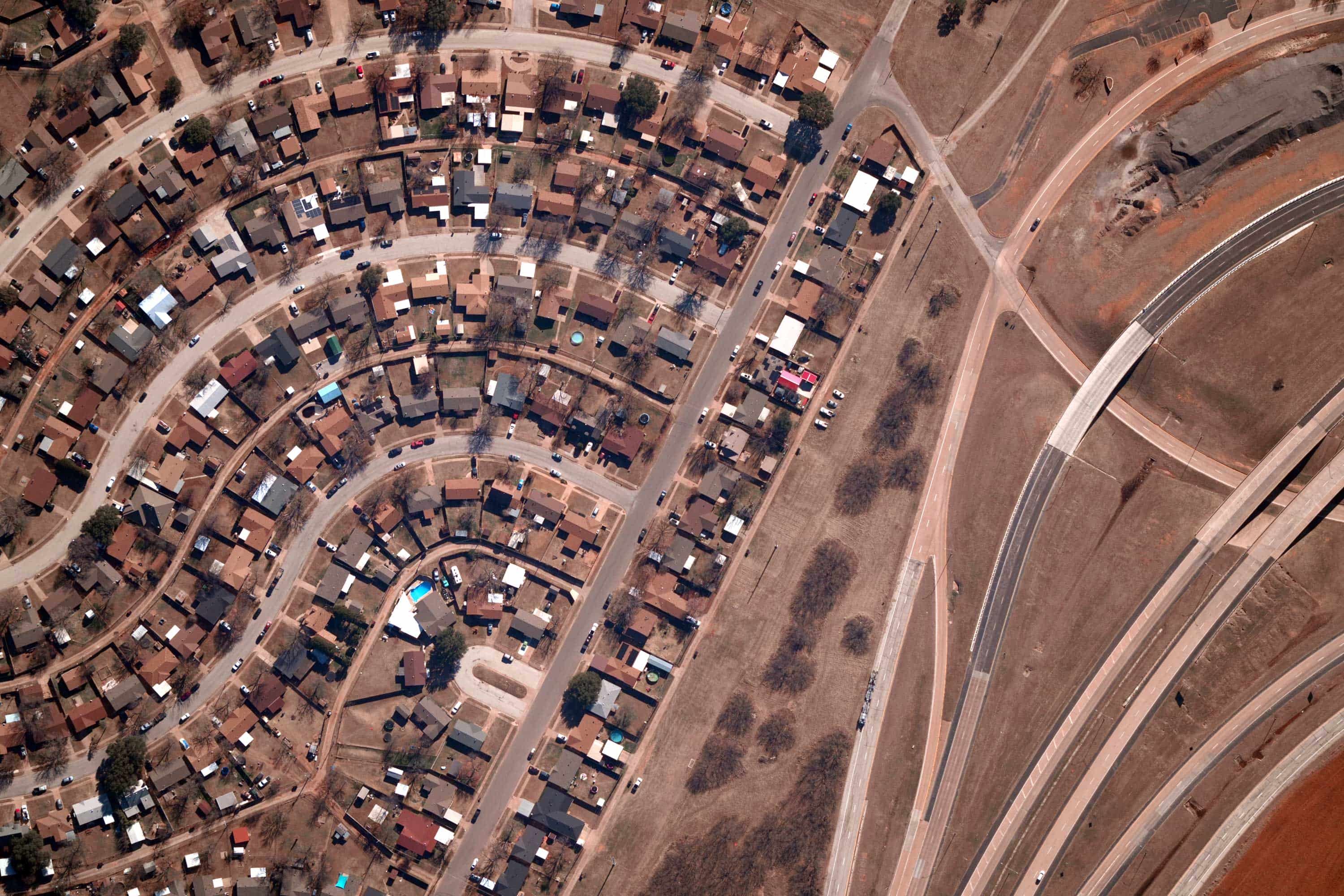 Aerial view of a residential neighborhood with curved streets and houses alongside open brown grassland and intersecting roads.