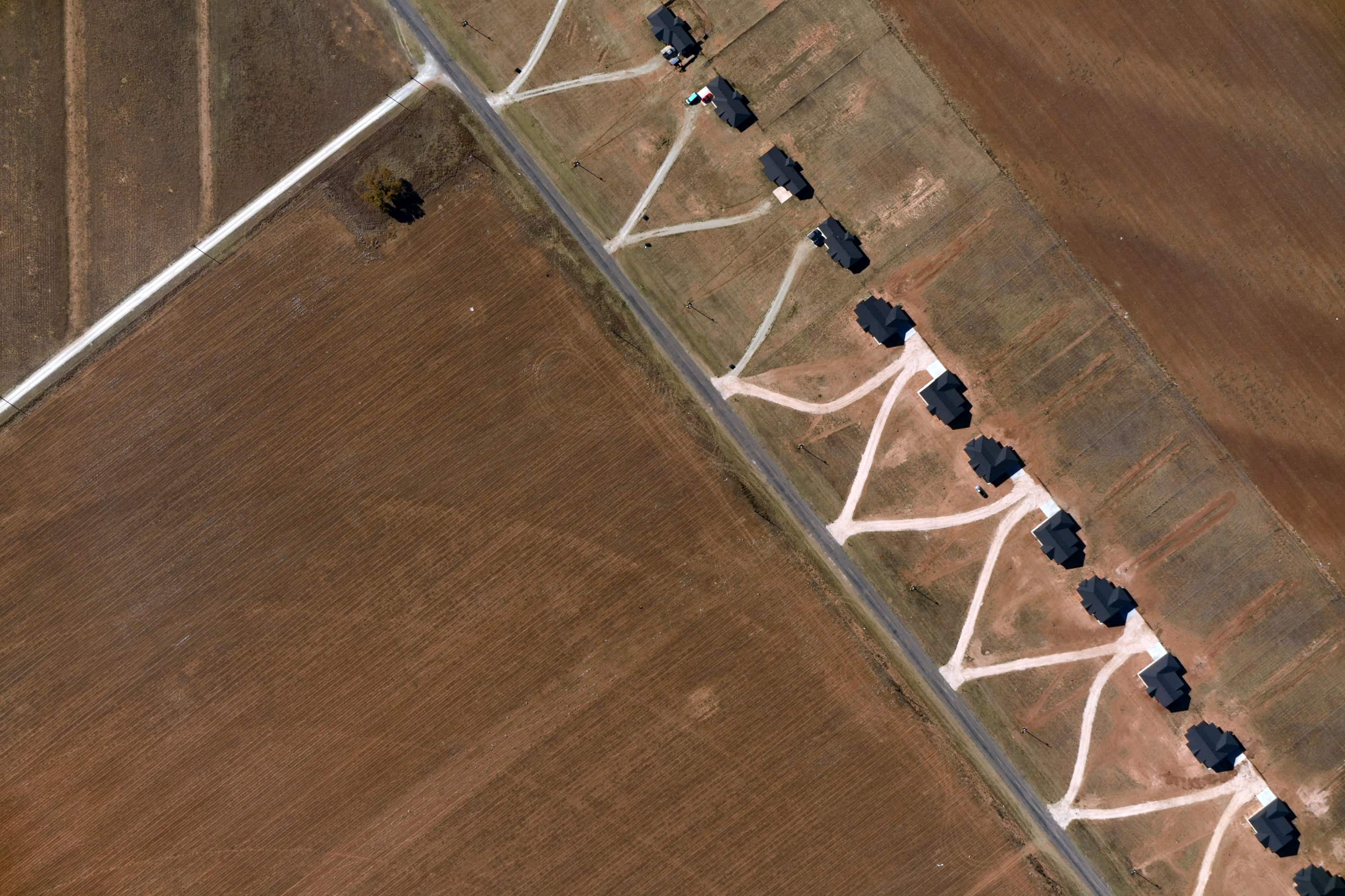 Aerial view of a rural road lined with houses having dirt driveways branching off from the road, surrounded by farmland fields.