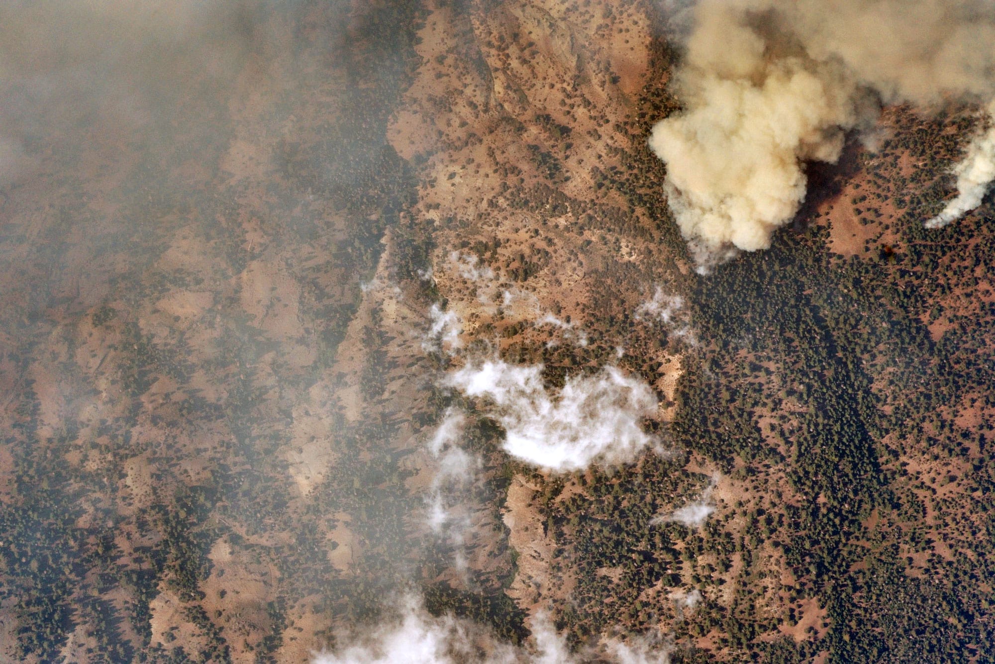 Aerial view of a wildfire with thick smoke rising above dry vegetation and scattered trees.