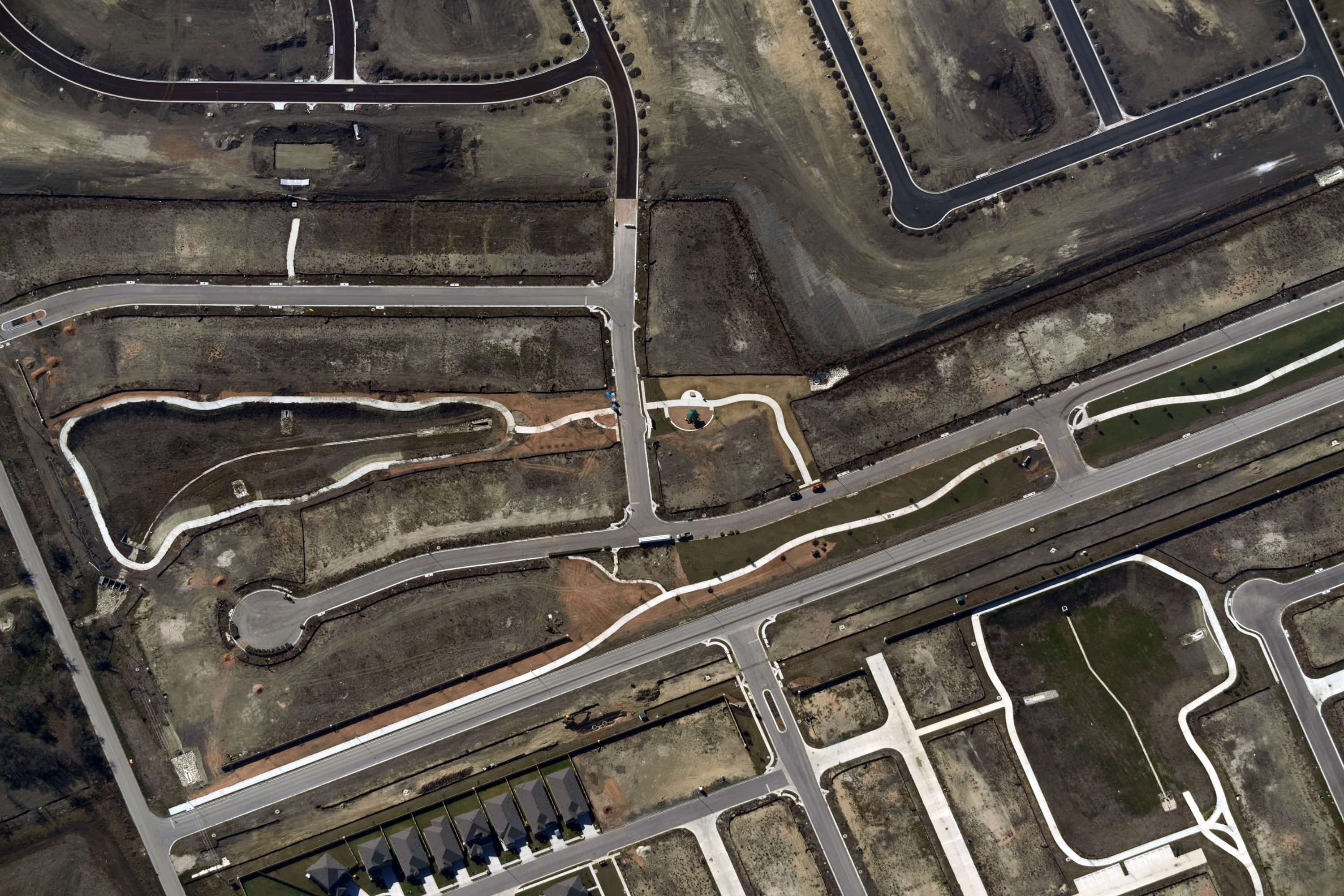 Aerial view of a partially developed suburban area with roads, sidewalks, curbs, and some houses constructed on empty plots of land.