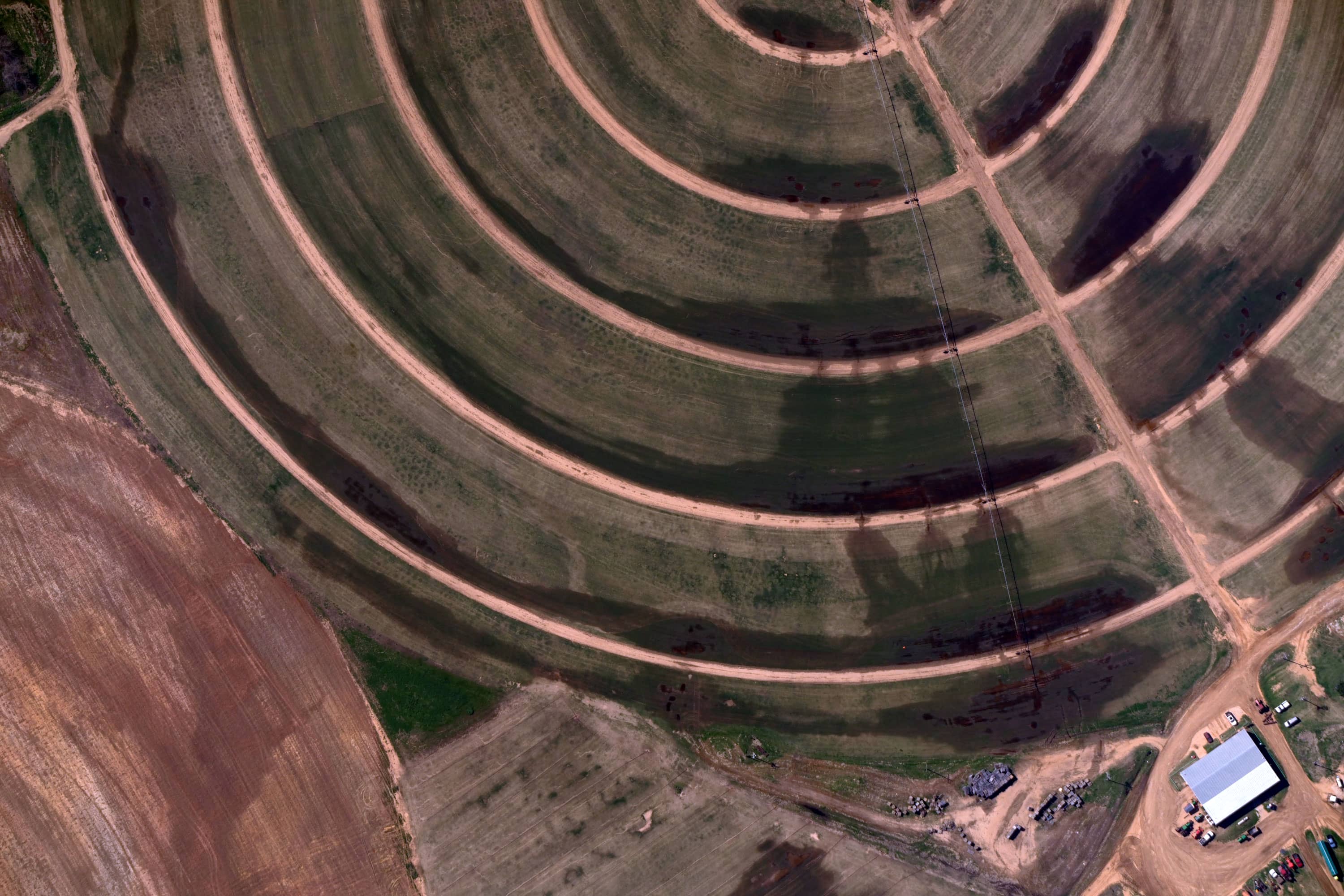 Aerial view of circular crop fields with dirt roads and a white-roofed building surrounded by parked vehicles.