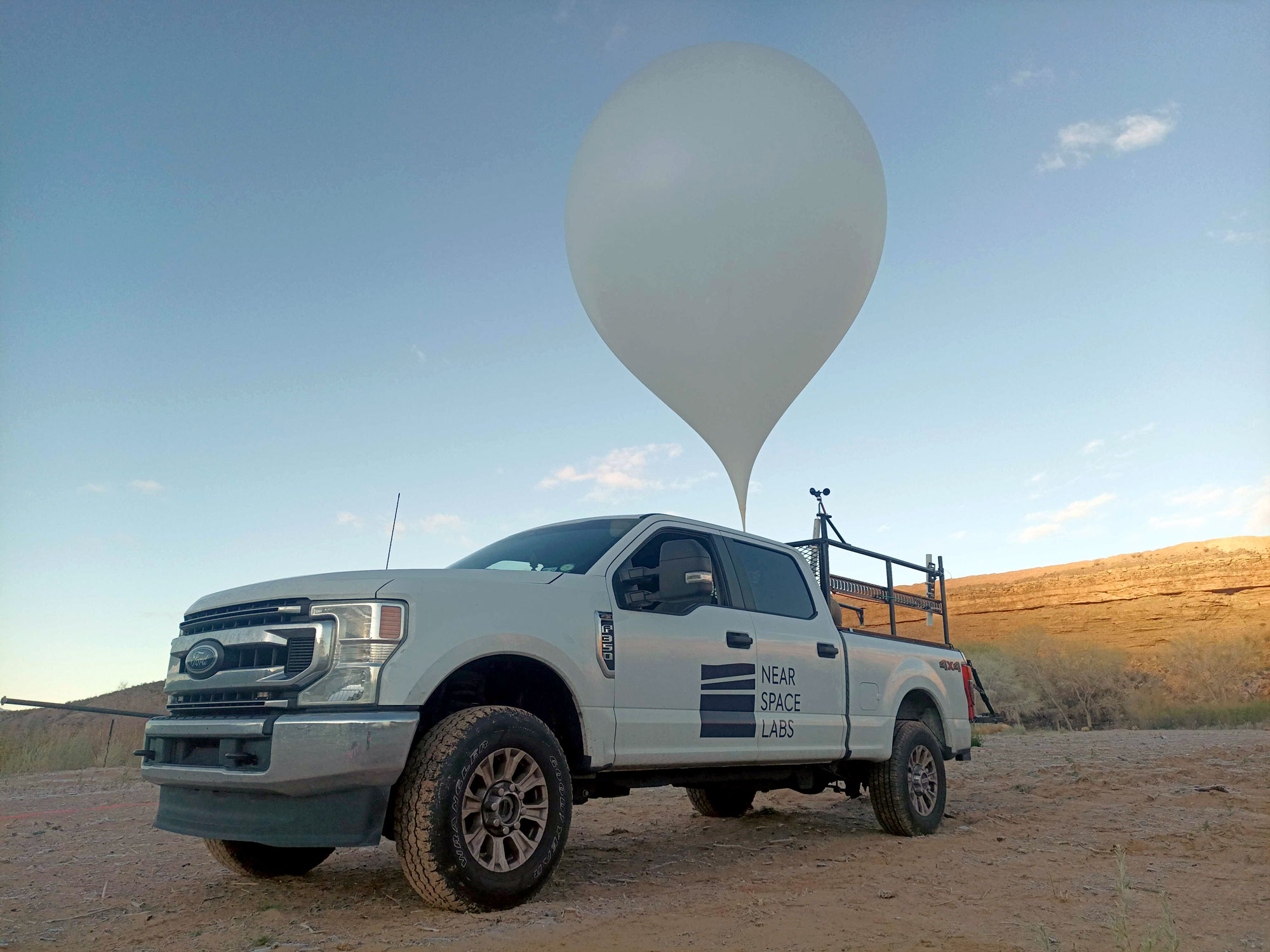 White Ford pickup truck with Near Space Labs logo carrying a large weather balloon on a rack, parked on a dirt road with hills in the background.
