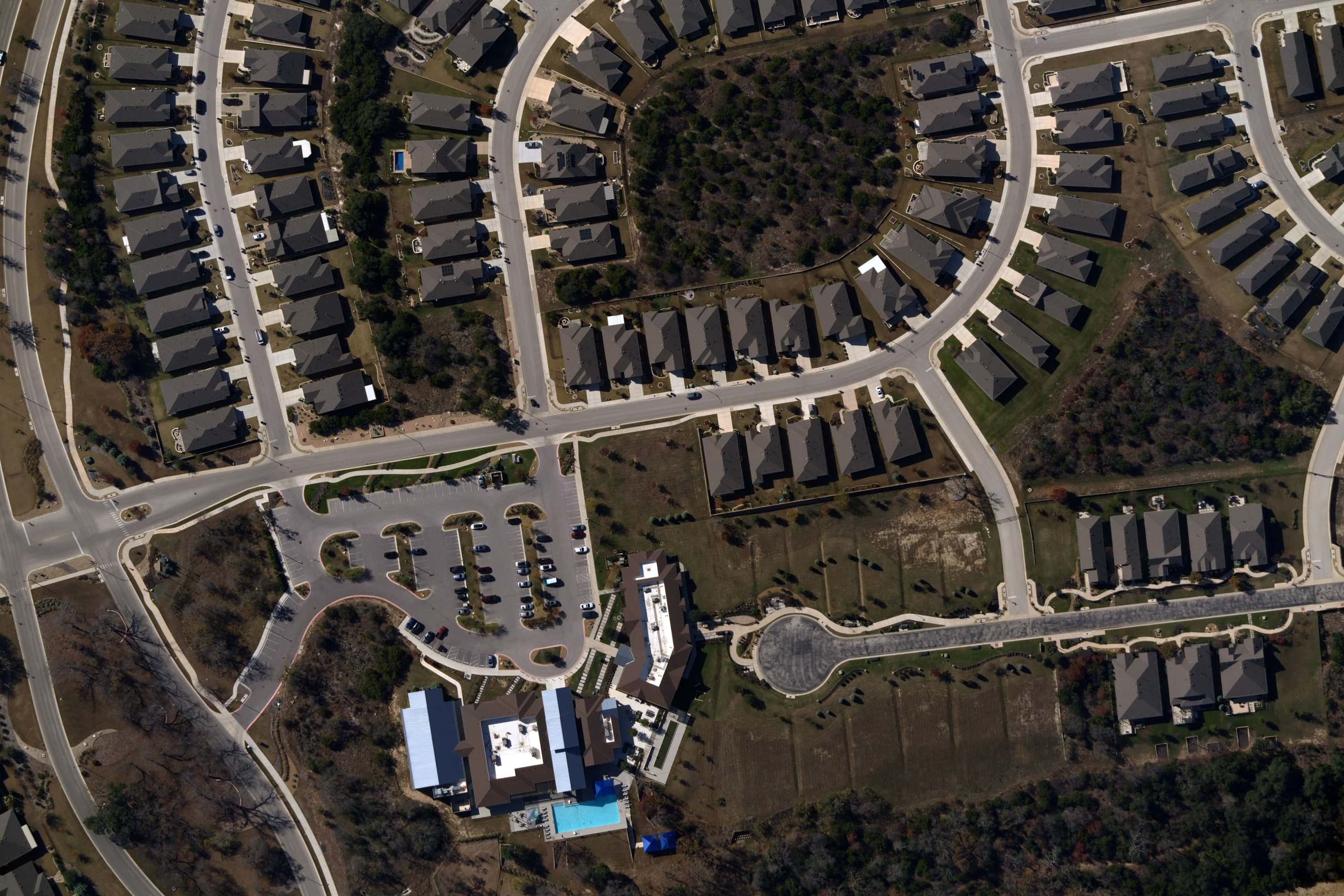 Aerial view of a suburban neighborhood with rows of houses, winding roads, parking lots, and a building with a swimming pool surrounded by trees.