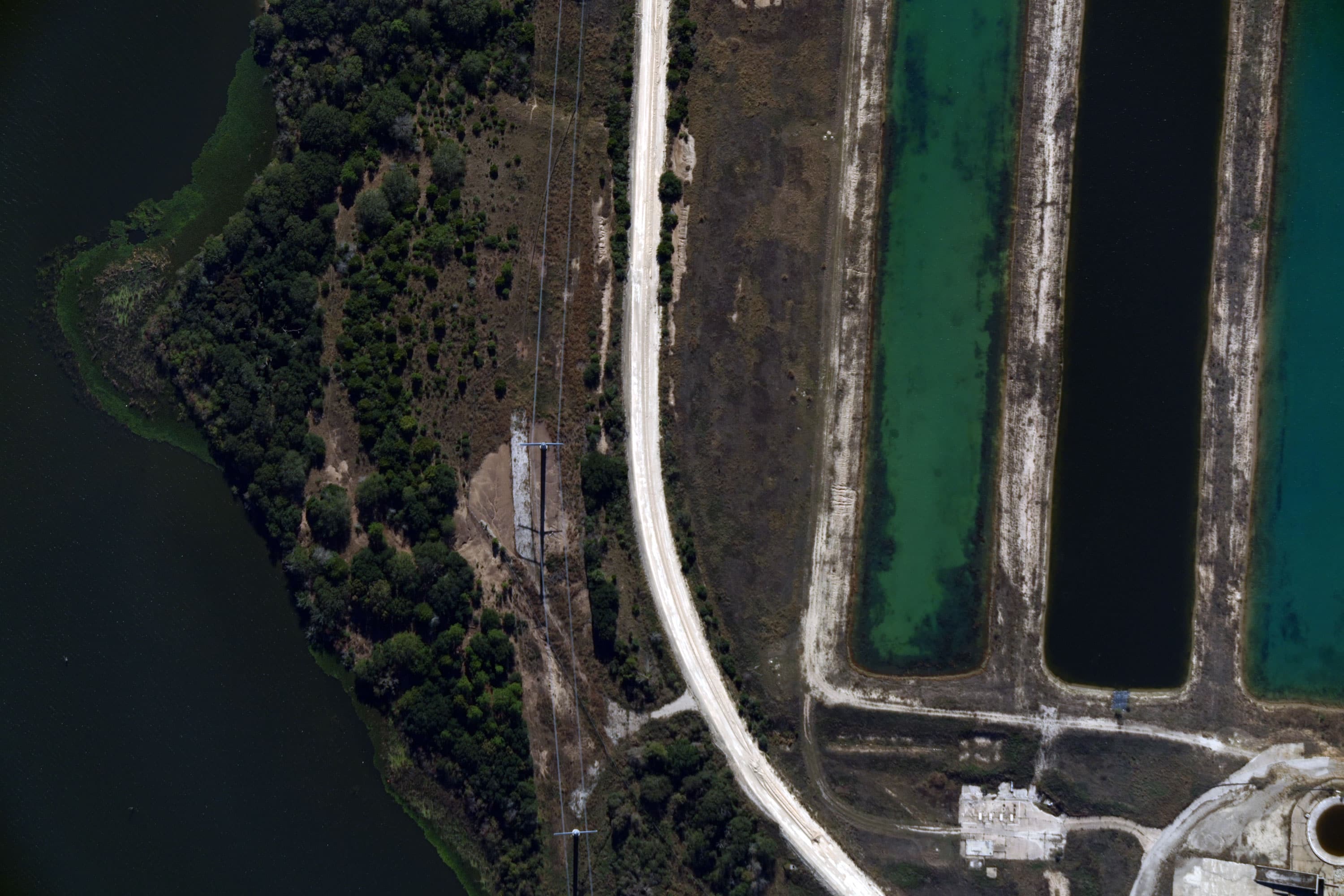 Aerial view of a curved dirt road flanked by forested land on one side and rectangular reservoirs on the other.