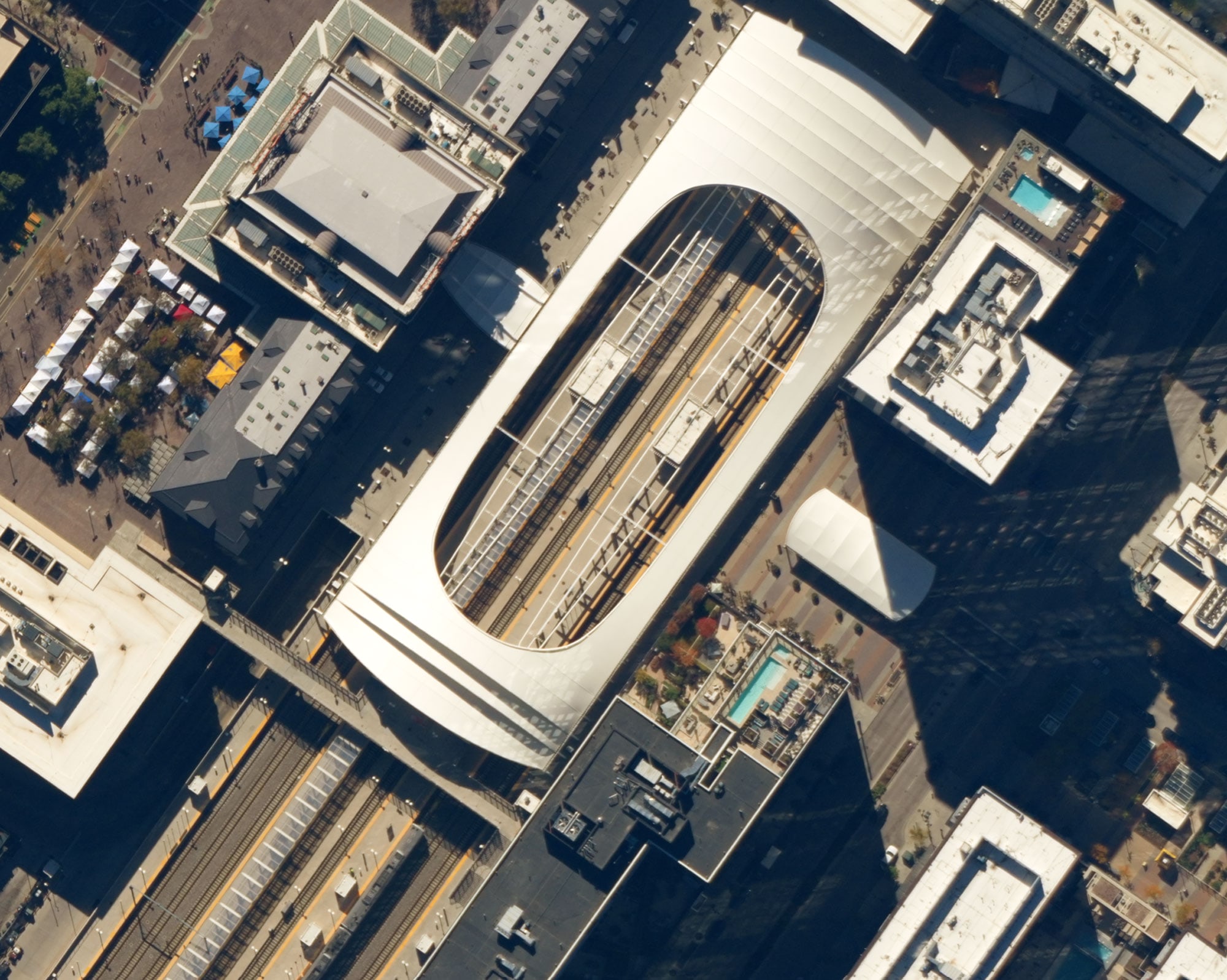 Aerial view of a large modern train station with a white curved roof and multiple tracks, surrounded by city buildings casting shadows.