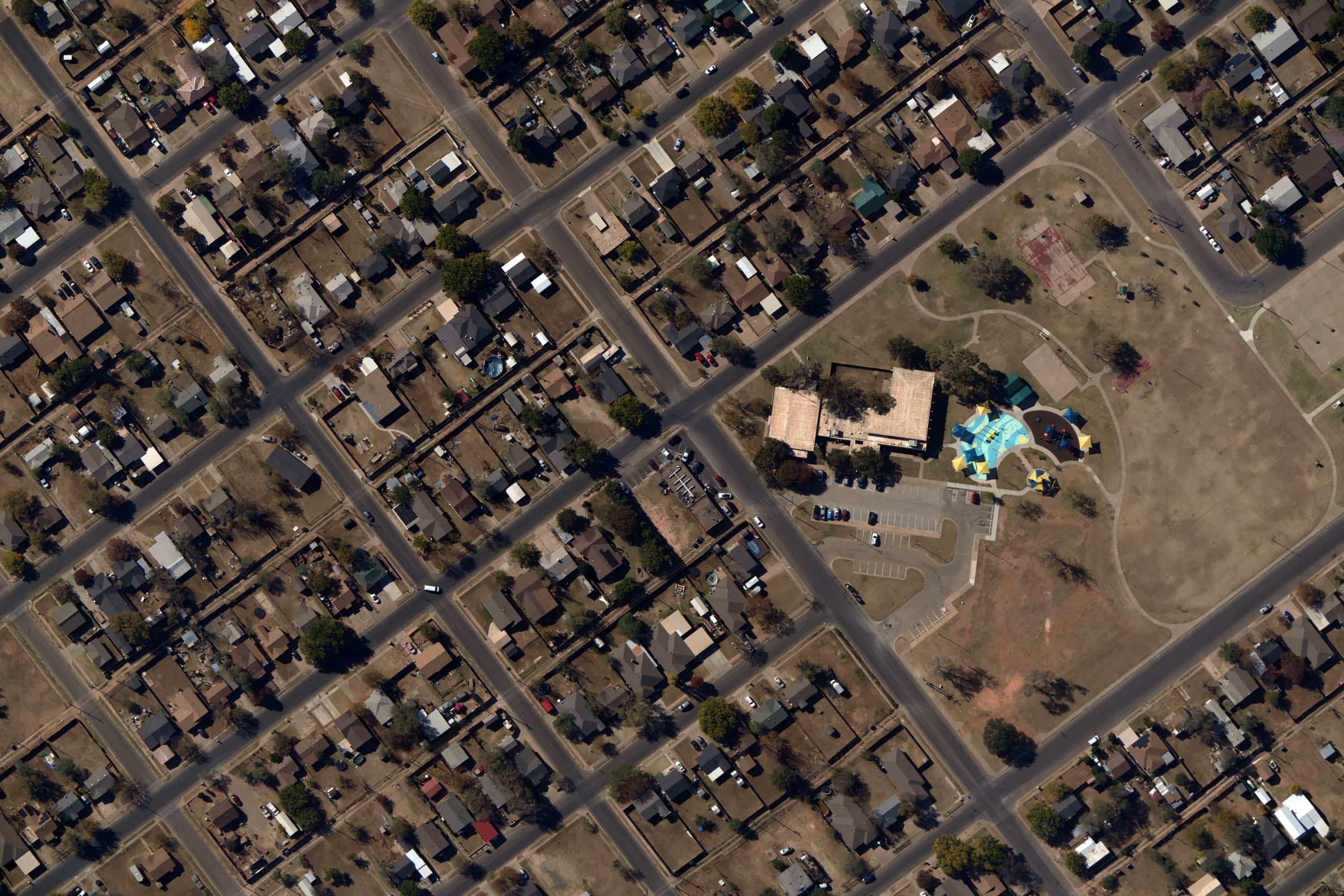 Aerial view of a suburban neighborhood with houses, streets, a parking lot, and a park area with colorful shade structures over playground equipment.