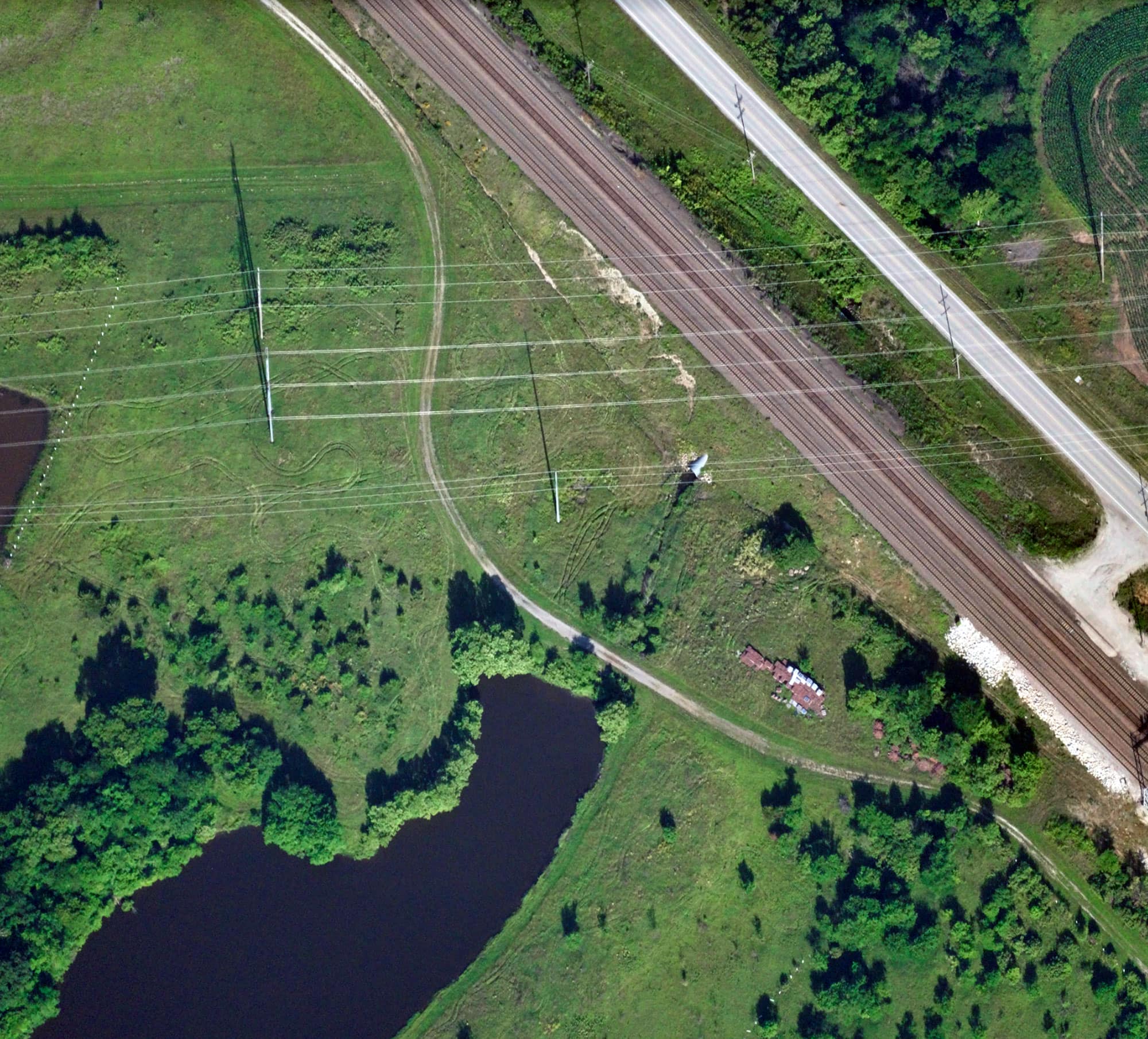 Aerial view of a green landscape with a pond, power lines, railroad tracks, a road, and patches of trees and grass.