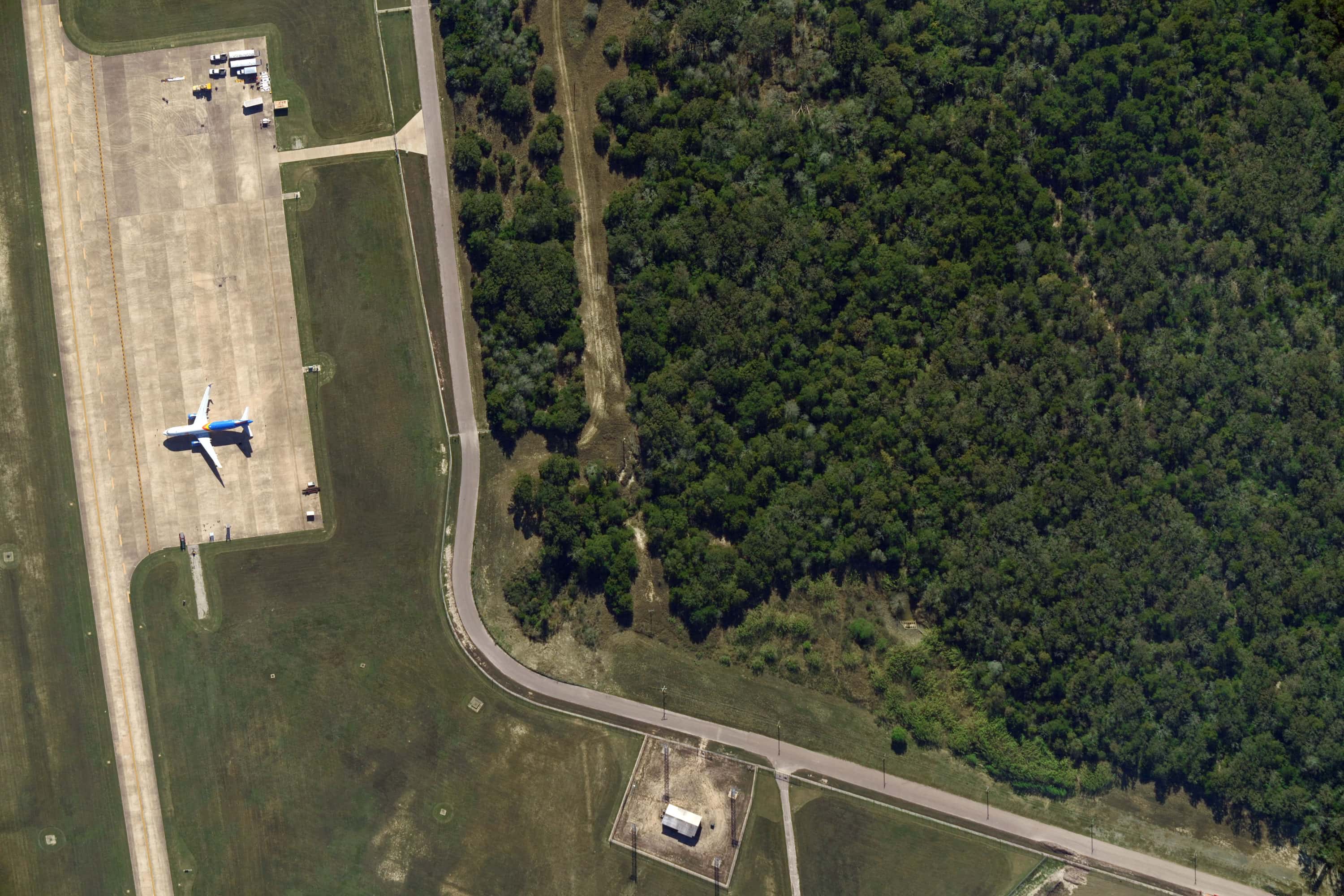Aerial view of a small airplane parked on a runway ramp next to a curved road and dense forest area.