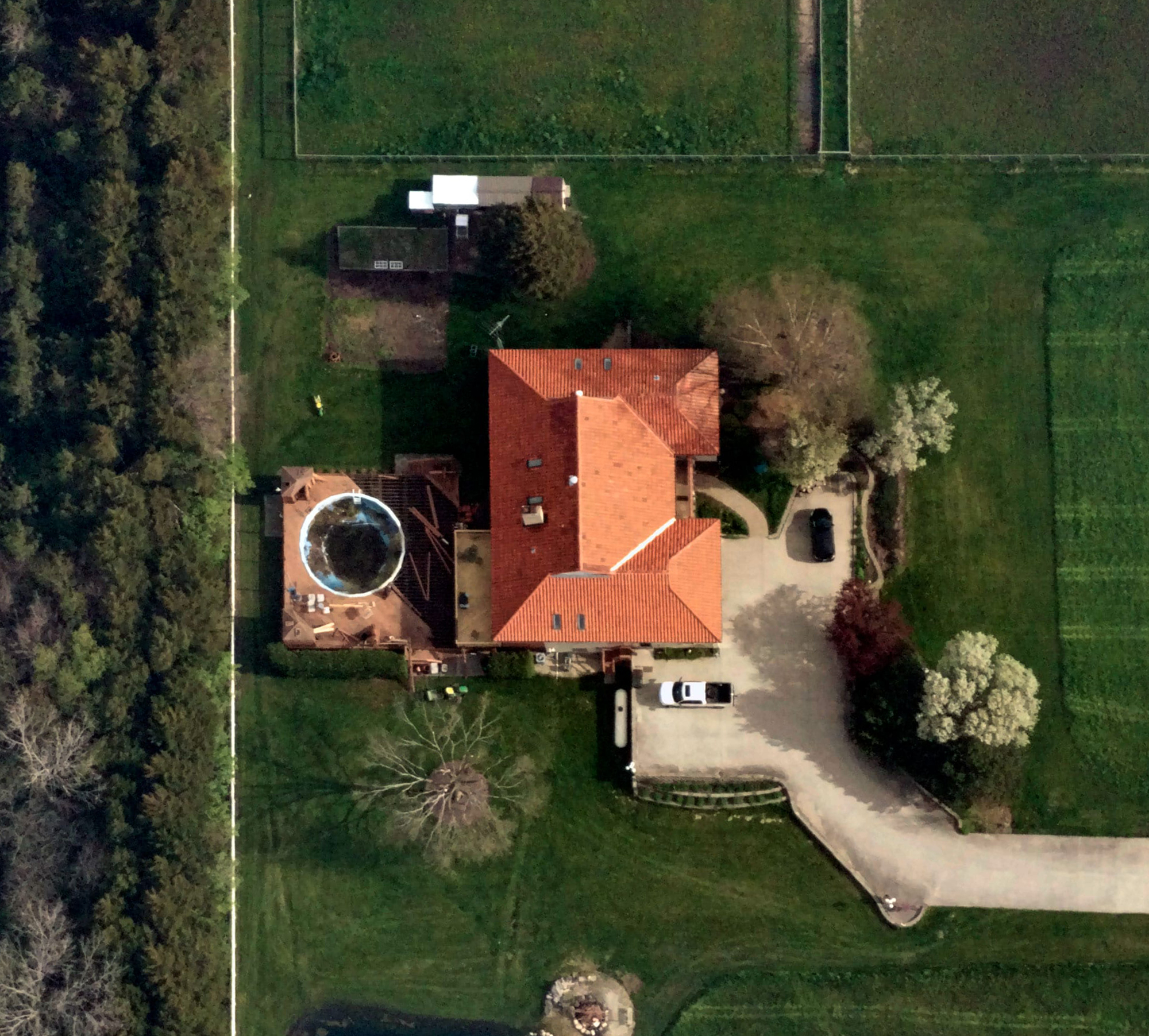 Aerial view of a house with a red tiled roof, a driveway with parked cars, surrounding green lawn, trees, and a circular above-ground pool.