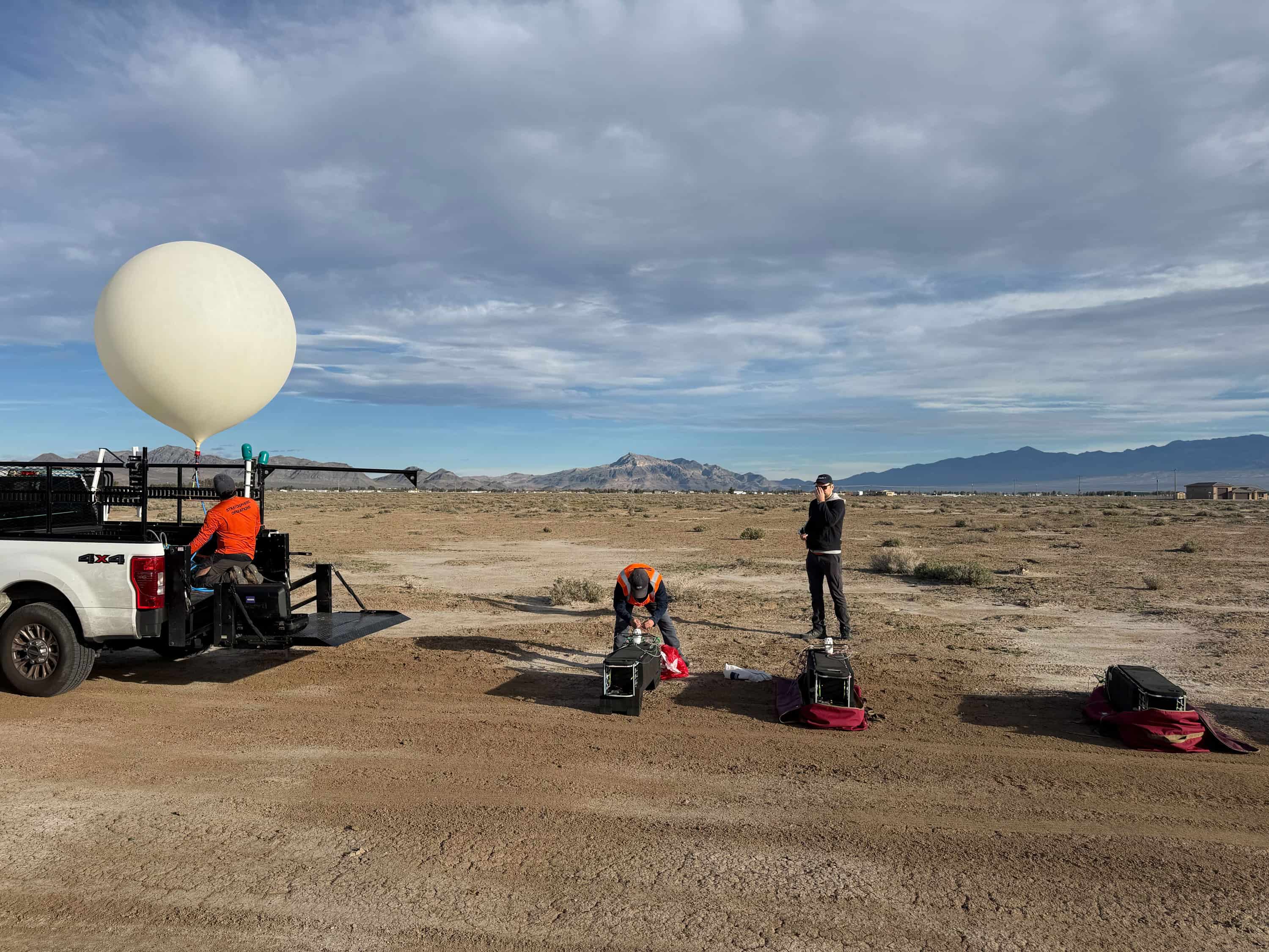 Three people preparing weather balloons and capture flight equipment in a desert area with mountains in the background.