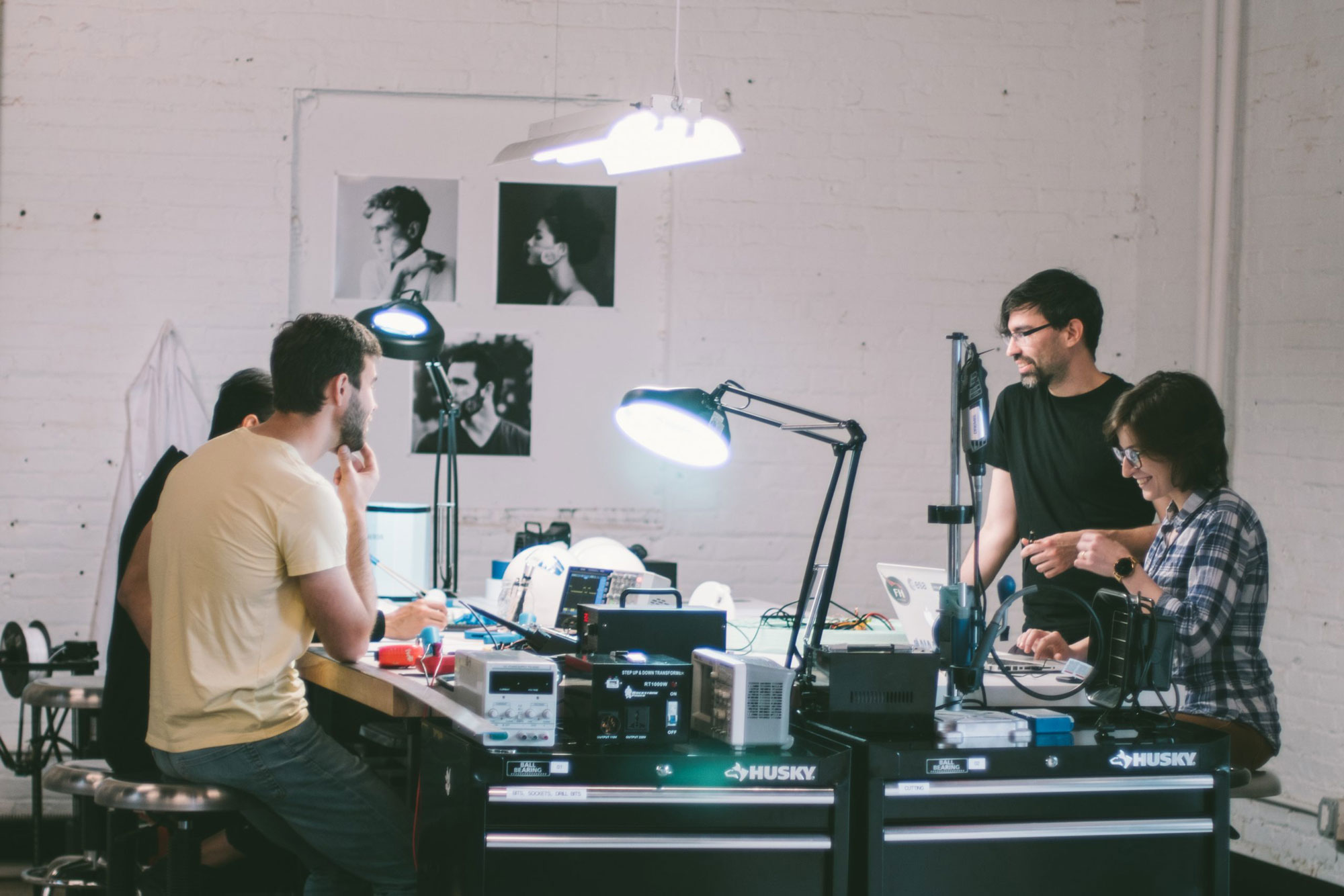 Group of four people working together on electronics and laptops in a bright workshop with tool cabinets and desk lamps.