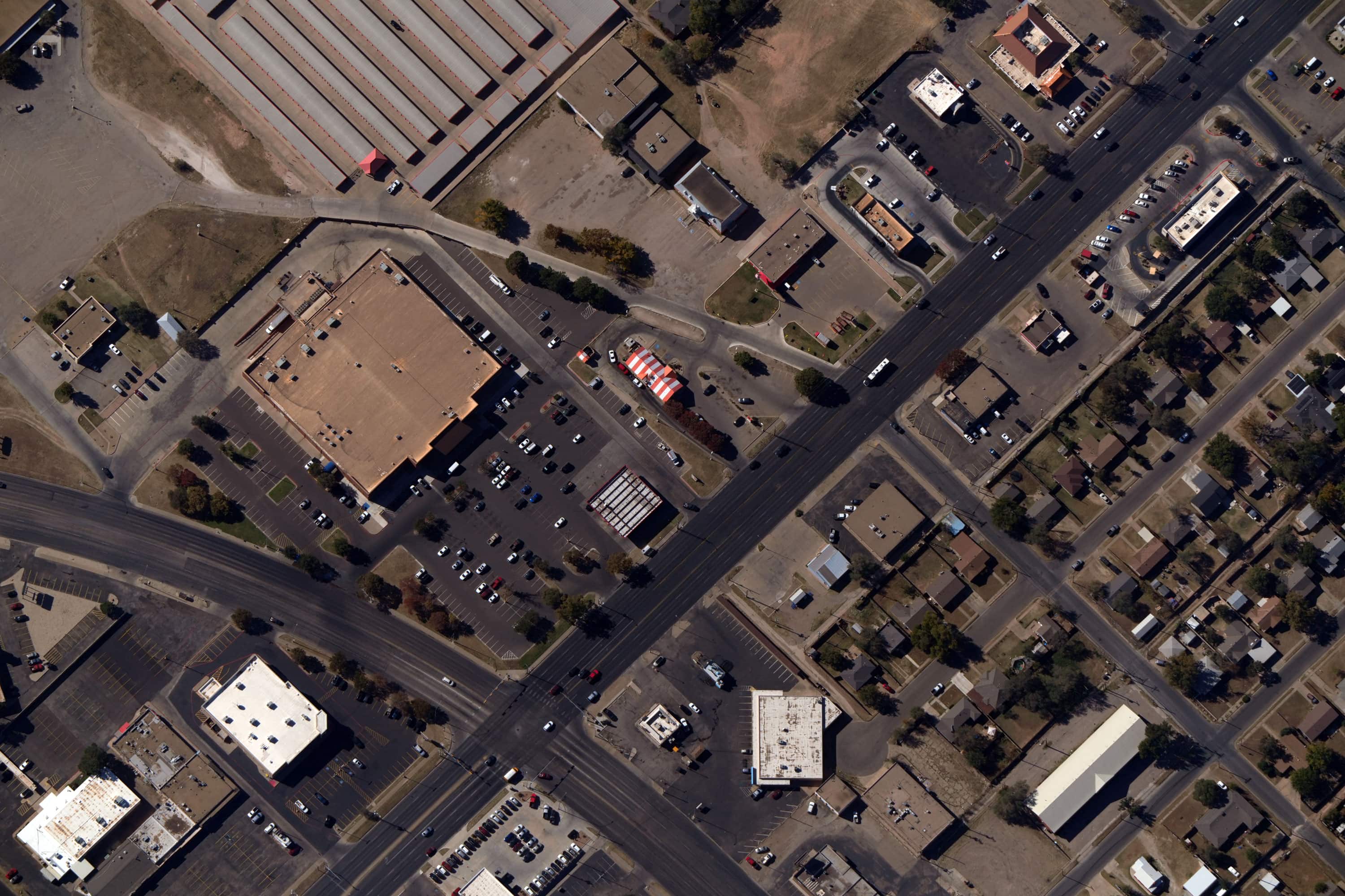 Aerial view of an urban intersection with commercial buildings, parking lots, cars, and nearby residential houses.
