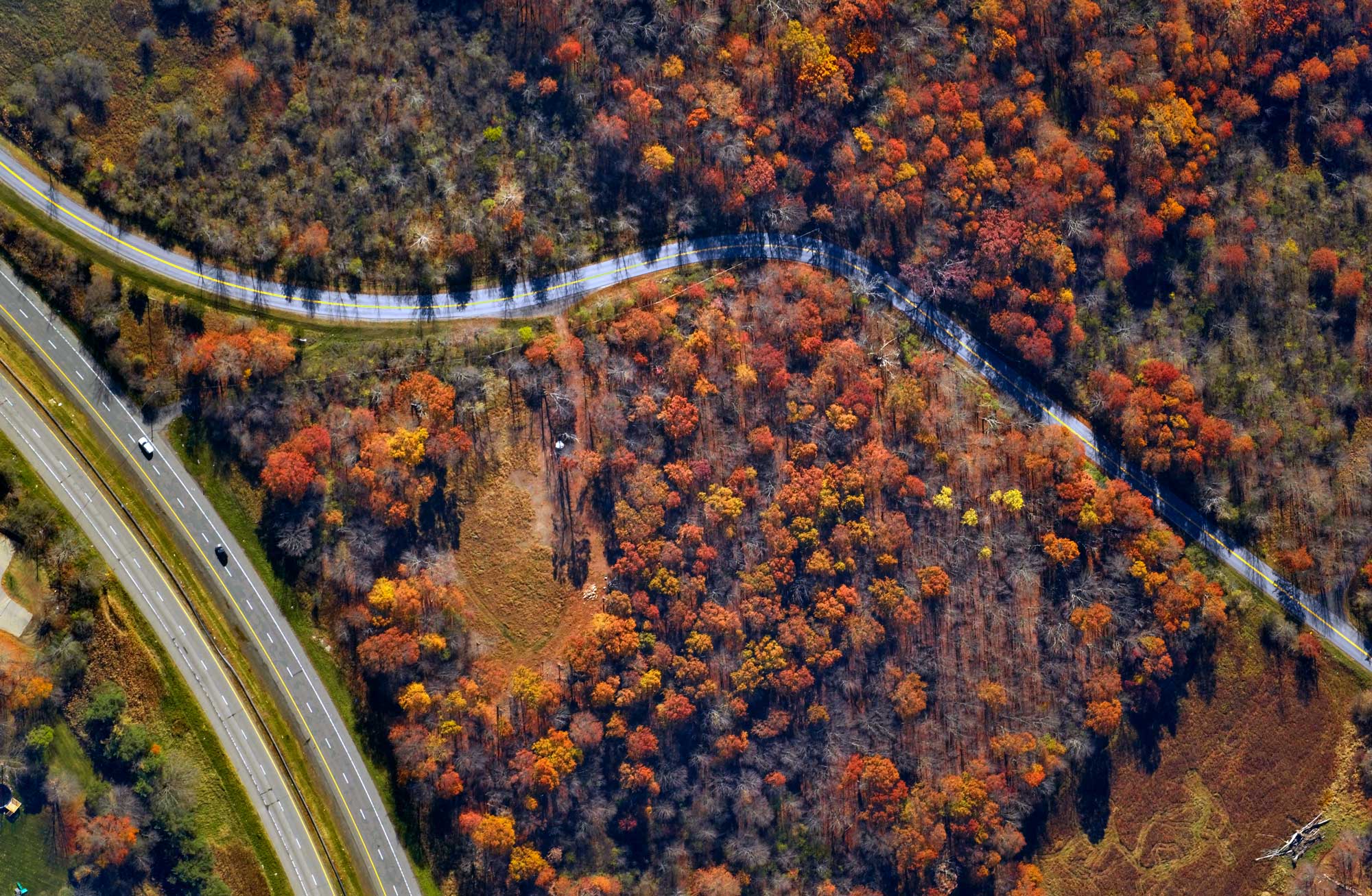Aerial view of a forest with colorful autumn foliage near a curving road and a highway with vehicles.