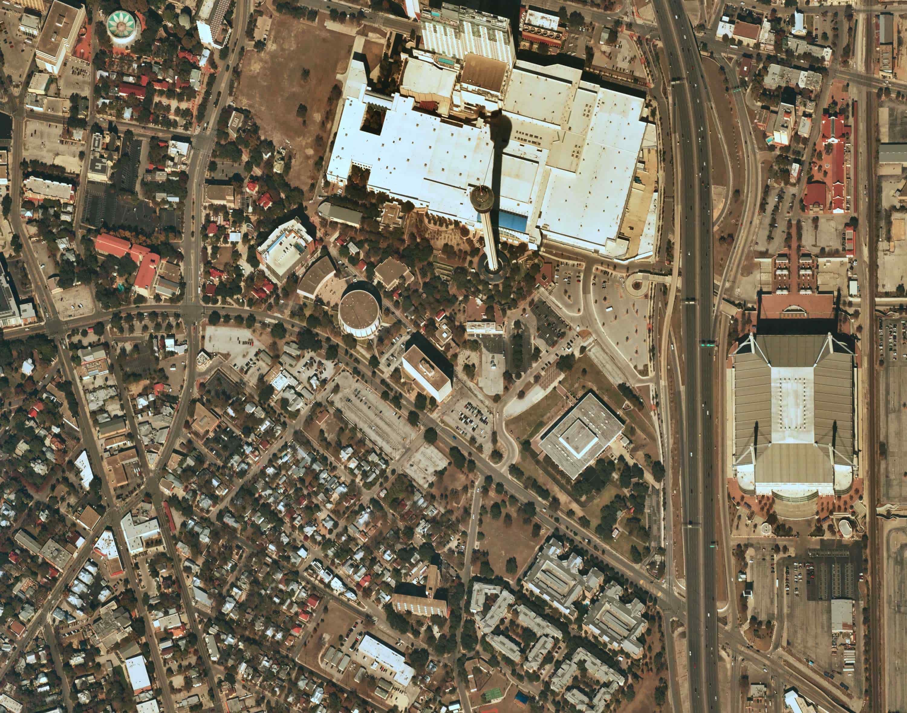 Aerial view of a city block featuring a tall observation tower casting a long shadow beside a large white-roofed building and nearby highway.