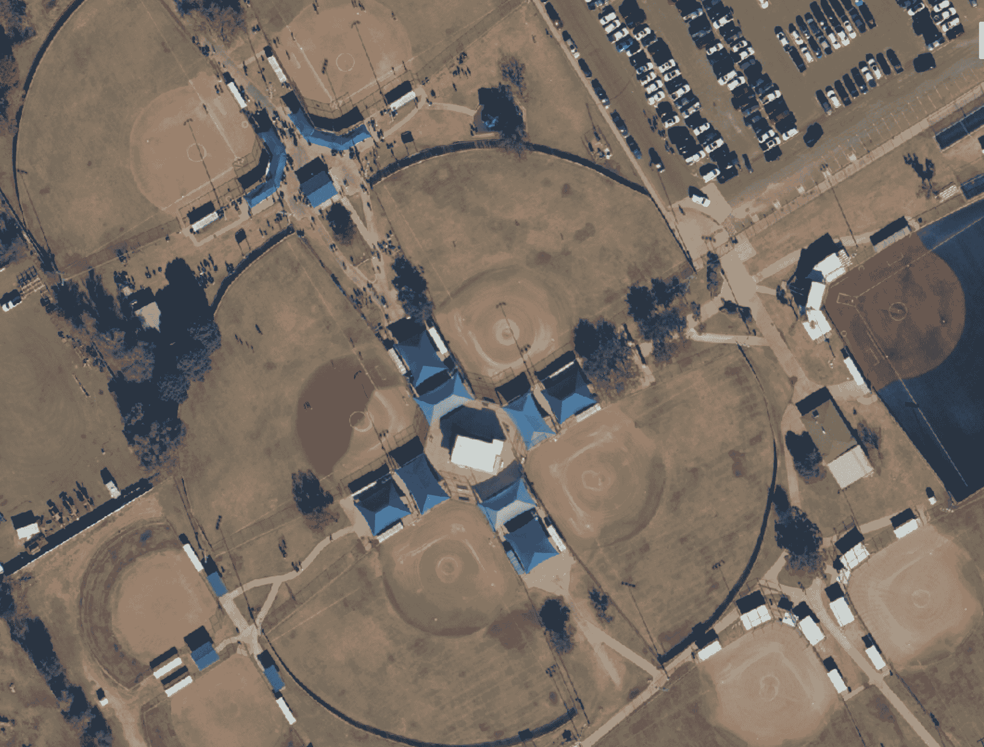 Detailed aerial view of a baseball complex with multiple baseball fields, blue tarps seen around the central fields, and parking lots with numerous cars.