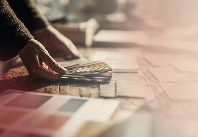 Person's hand selecting a color from a fan deck on a wooden table surrounded by color samples.