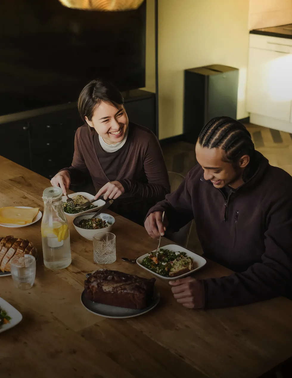 Two people sitting at a wooden table smiling and eating healthy meals with bread and lemon water, with text overlay 'Built around you. Nutrition Plans.'