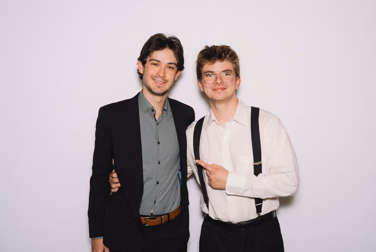 Deux jeunes hommes souriants, l'un en veste noire et chemise grise, l'autre en chemise blanche avec des bretelles, pointant vers son ami.