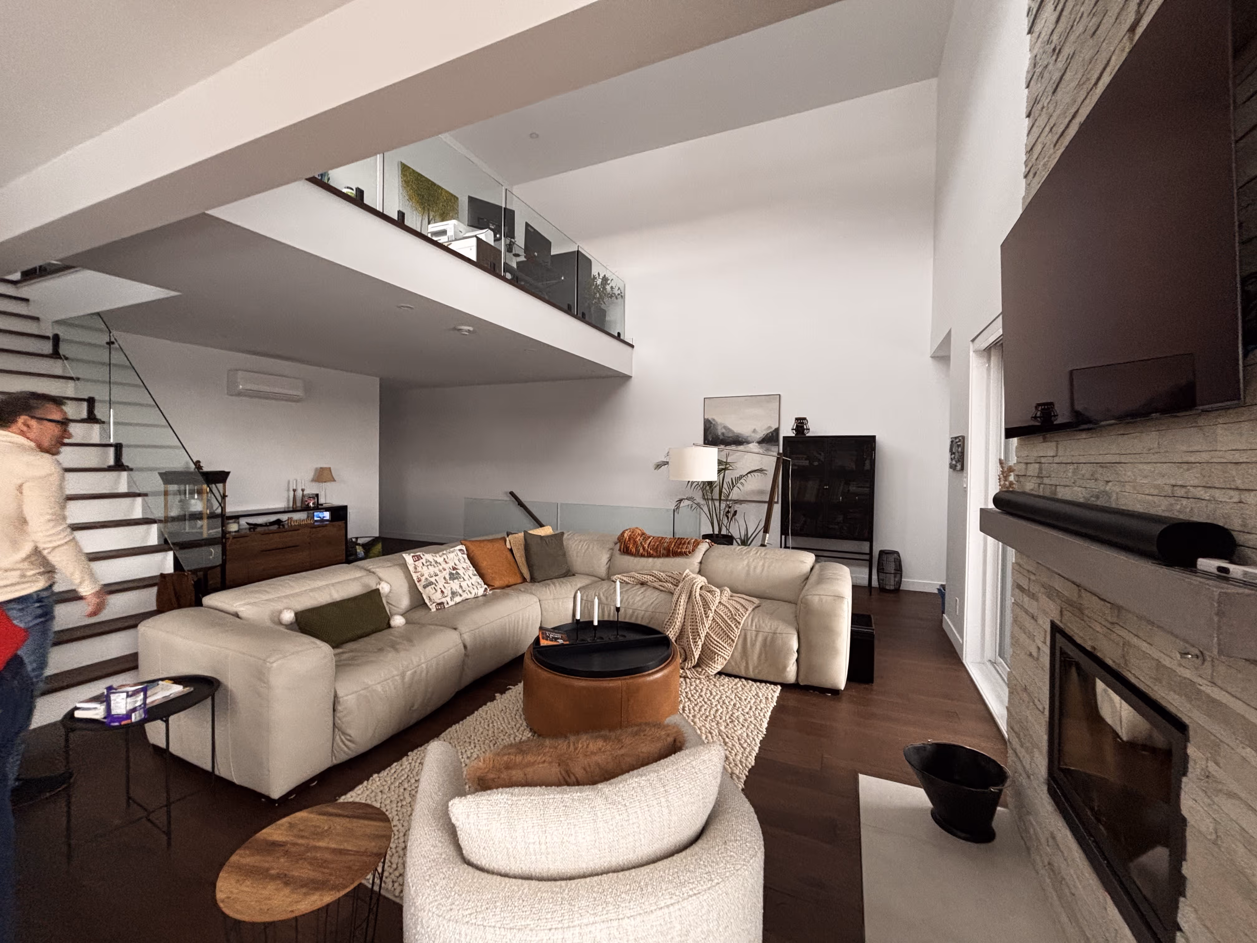 Salon moderne avec canapé en cuir beige, fauteuil rond, table basse, télévision murale au-dessus d'une cheminée en pierre et escalier menant à un étage supérieur.