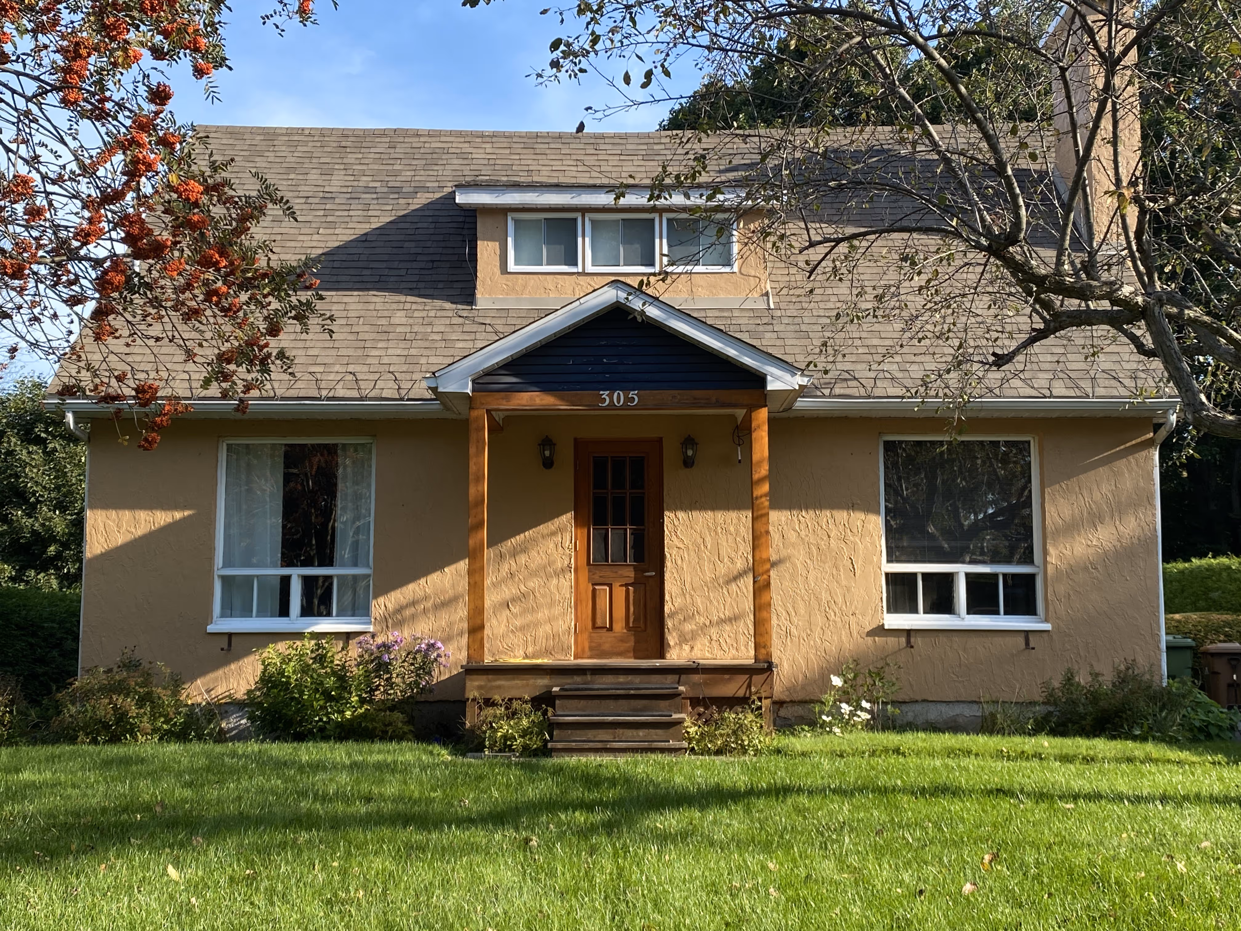 Façade d'une maison beige avec un porche en bois, deux fenêtres blanches et un numéro 305 au-dessus de la porte.