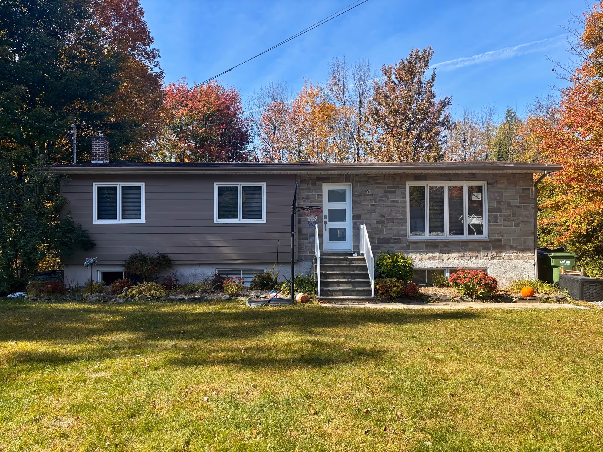 Maison unifamiliale en brique et bois avec une petite pelouse à l’avant, entourée d’arbres aux feuilles d'automne colorées.