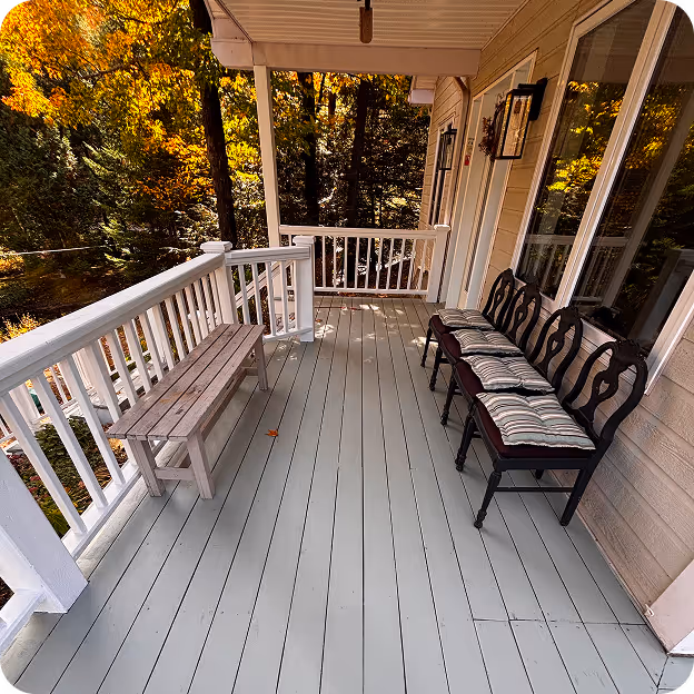 Porche en bois peint gris avec bancs en bois et chaises à coussins, entouré d'arbres aux feuilles d'automne.