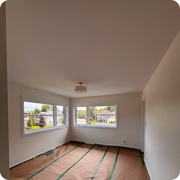 Chambre vide avec grandes fenêtres d'angle offrant une vue sur un quartier résidentiel, sol couvert de papier protecteur marron avec bandes vertes.