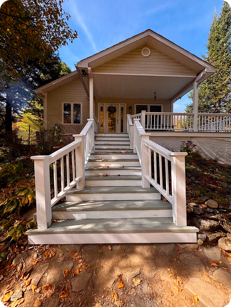 Escalier blanc avec rampe menant à un porche couvert d'une maison entourée d'arbres en automne sous un ciel bleu clair.