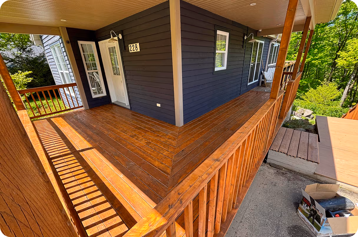 Porche en bois autour d'une maison avec façade bleue, porte blanche et numéro 228, entouré d'arbres verts.
