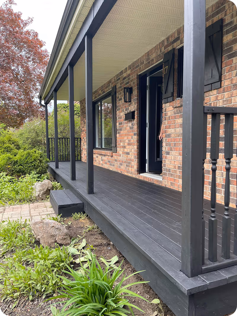 Porche noir d'une maison en brique avec des plantes vertes et un arbre aux feuilles rouges à gauche.