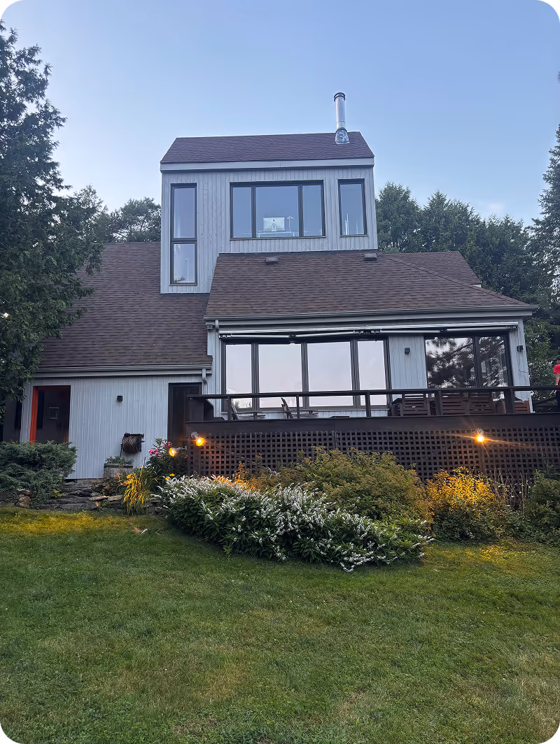 Maison à deux étages avec bardage gris, nombreuses fenêtres, terrasse en bois et jardin bien entretenu au crépuscule.