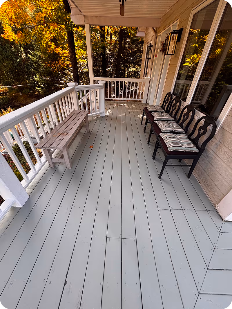 Porche en bois peint gris avec banc rustique à gauche et trois chaises noires garnies de coussins à rayures à droite, entouré d'arbres à feuilles d'automne.