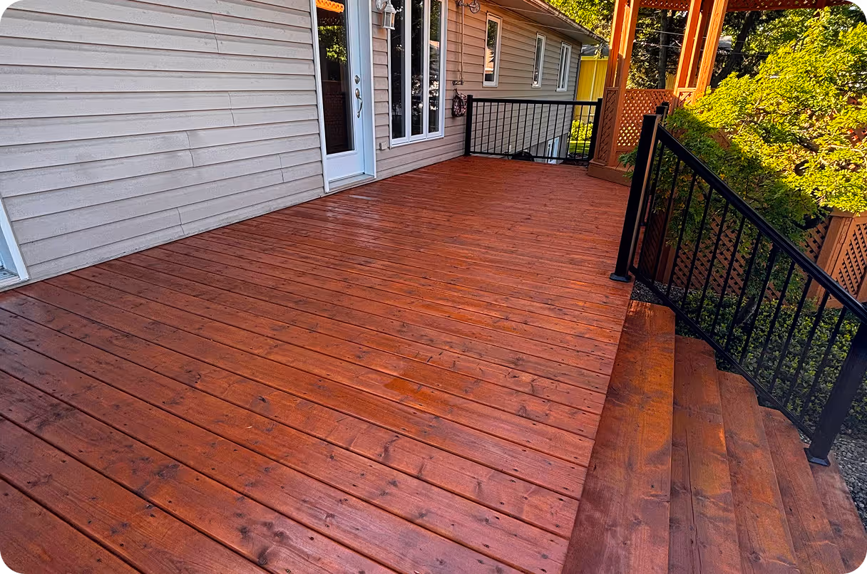 Grand patio en bois teinté rouge avec rambardes noires et escaliers, attenant à une maison avec porte blanche et fenêtres.