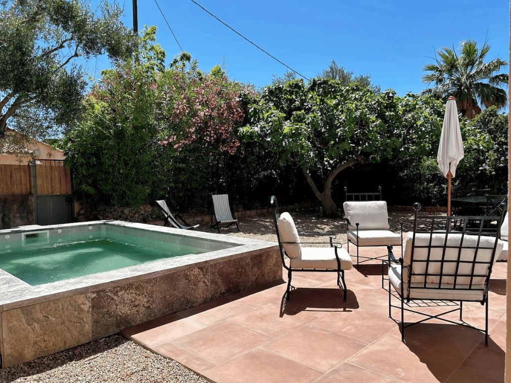 Coin terrasse avec chaises métalliques blanches autour d'une petite piscine sur une surface carrelée, entourée de végétation sous un ciel bleu clair.