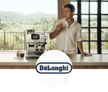 Un homme en chemise blanche tenant un verre de café devant une machine à café DeLonghi sur un comptoir avec une vue sur un paysage verdoyant.