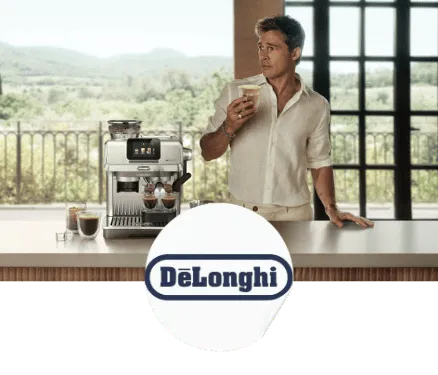 Un homme en chemise blanche tenant un verre de café devant une machine à café DeLonghi sur un comptoir avec une vue sur un paysage verdoyant.