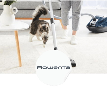 Une personne nettoie un tapis clair avec un aspirateur Rowenta pendant qu'un chat marche à côté.