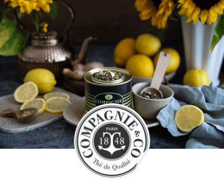 Pot de thé noir Compagnie & Co entouré de citrons, cuillères en bois et tournesols sur une table.