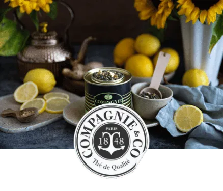 Pot de thé noir Compagnie & Co entouré de citrons, cuillères en bois et tournesols sur une table.