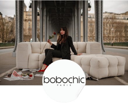 Femme en tailleur noir et chaussures rouges tenant une rose assise sur un canapé blanc en extérieur sous une structure métallique.