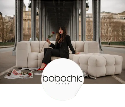 Femme en tailleur noir et chaussures rouges tenant une rose assise sur un canapé blanc en extérieur sous une structure métallique.