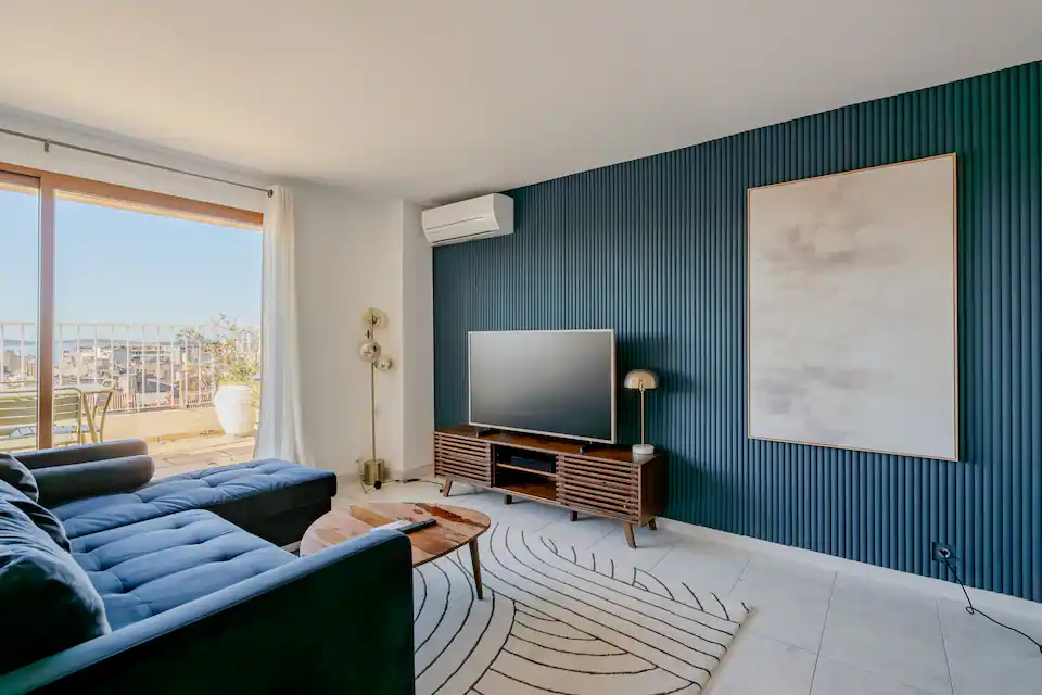 Salon moderne avec canapé bleu, table basse en bois, meuble TV en bois, mur bleu cannelé et grandes baies vitrées donnant sur un balcon avec mobilier extérieur.