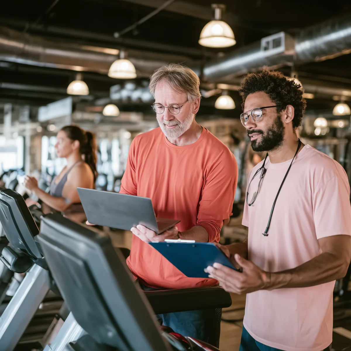 Zwei Männer besprechen Trainingsdaten auf einem Laptop in einem Fitnessstudio neben Laufbändern.