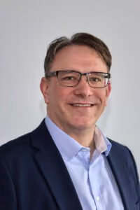 Mann mittleren Alters mit Brille und dunklem Sakko lächelnd vor hellem Hintergrund.
